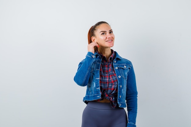 Jonge vrouw trekt oorlel in geruit hemd, jas, broek en ziet er vrolijk uit. vooraanzicht.