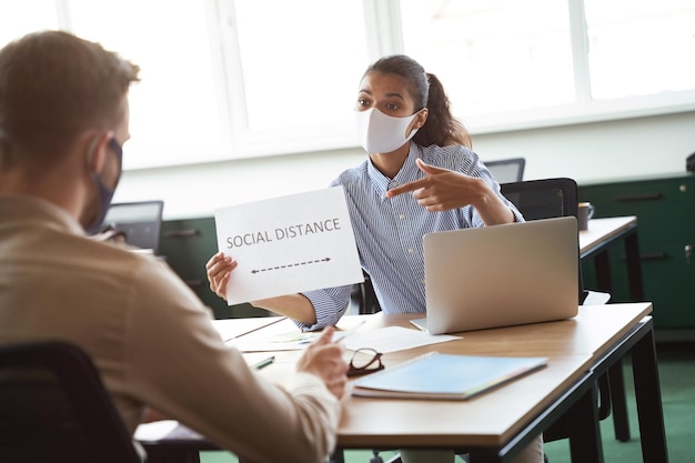 Jonge vrouw van gemengd ras die een beschermend gezichtsmasker draagt en papier toont met tekst sociale afstand tot haar