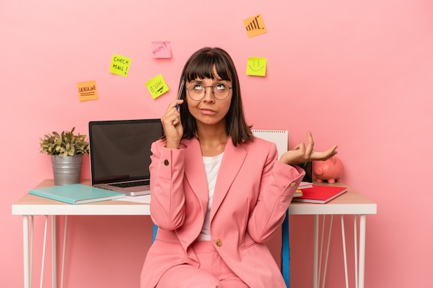 Jonge vrouw van gemengd ras die een examen voorbereidt in de kamer met een pen geïsoleerd op roze achtergrond