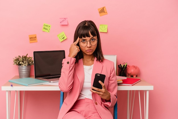 Jonge vrouw van gemengd ras die een examen voorbereidt voor de universiteit met een mobiele telefoon geïsoleerd op een roze achtergrond die naar de tempel wijst met de vinger denken gericht op een taak