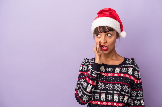 Jonge vrouw van gemengd ras die Kerstmis viert, geïsoleerd op een paarse achtergrond, zegt een geheim heet remnieuws en kijkt opzij