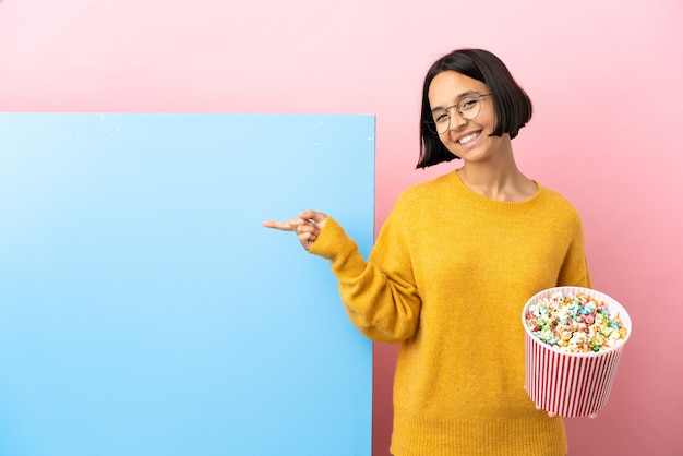 Jonge vrouw van gemengd ras die popcorn vasthoudt met een grote banner over een geïsoleerde achtergrond die met de vinger naar de zijkant wijst en een product presenteert