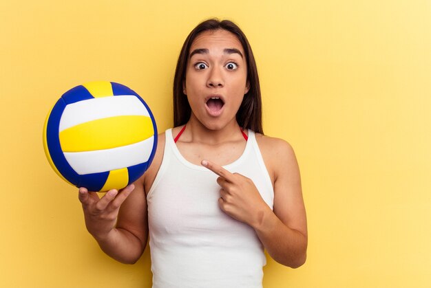 Jonge vrouw van gemengd ras die volleybal speelt op het strand geïsoleerd op een gele achtergrond die naar de zijkant wijst