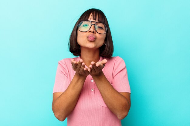 Jonge vrouw van gemengd ras geïsoleerd op blauwe achtergrond die lippen vouwt en handpalmen vasthoudt om luchtkus te sturen.