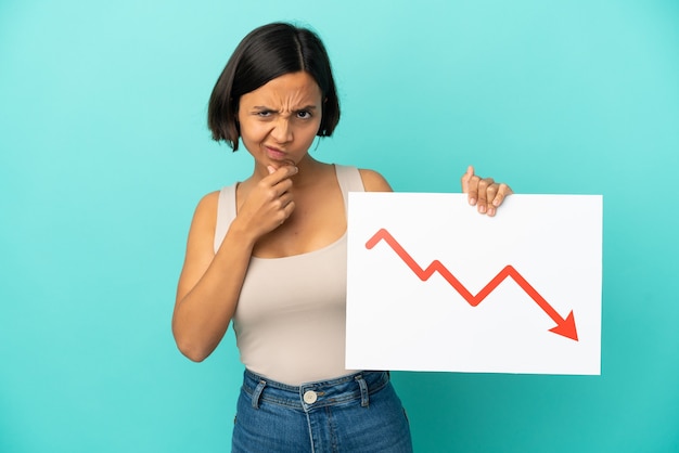 Jonge vrouw van gemengd ras geïsoleerd op een blauwe achtergrond met een bord met een dalend statistiekpijlsymbool en denkend