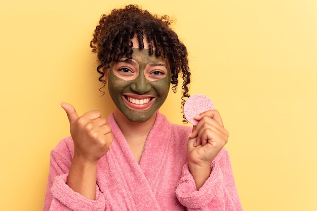 Jonge vrouw van gemengd ras, gekleed in een badjas met een make-up remover spons geïsoleerd op een gele achtergrond glimlachend en duim omhoog