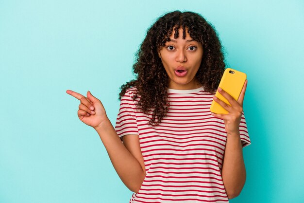 Jonge vrouw van gemengd ras met een mobiele telefoon geïsoleerd op een blauwe achtergrond die naar de zijkant wijst