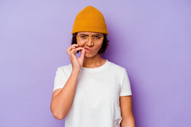 Jonge vrouw van gemengd ras met een wollen muts geïsoleerd op een paarse achtergrond met vingers op de lippen die een geheim bewaren.