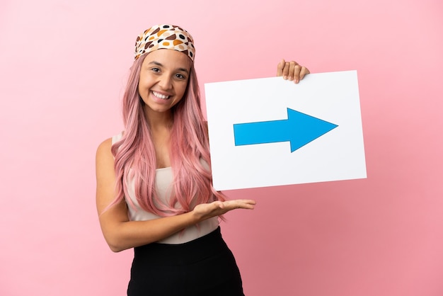 Jonge vrouw van gemengd ras met roze haar geïsoleerd op roze achtergrond met een bordje met pijlsymbool met gelukkige uitdrukking