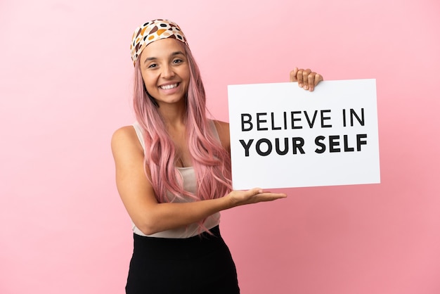 Jonge vrouw van gemengd ras met roze haar geïsoleerd op roze achtergrond met een bordje met tekst Believe In Your Self en erop wijzend