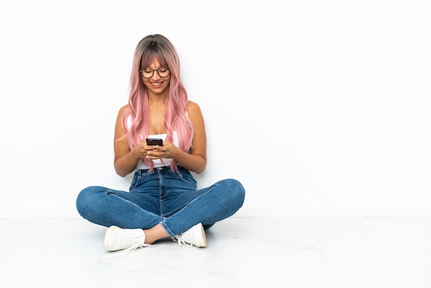 Jonge vrouw van gemengd ras met roze haar zittend op de vloer geïsoleerd op een witte achtergrond die een bericht verzendt met de mobiel