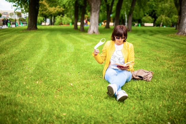 Jonge vrouw van middelbare leeftijd in vrijetijdskleding zet op een wit medisch masker zittend op de vloer in een zomers zonnig park