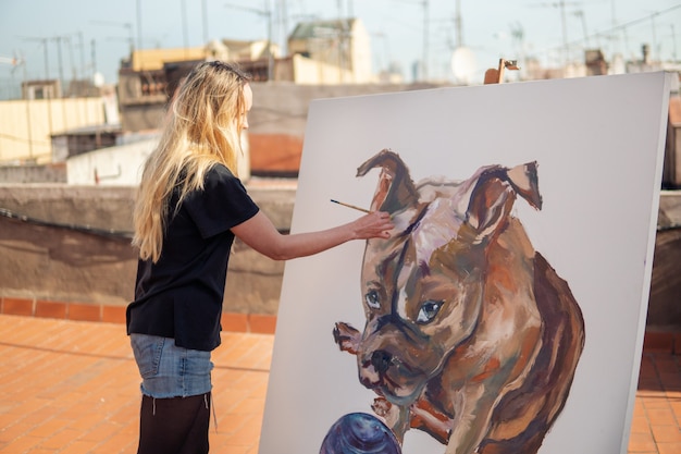 Jonge vrouw verf kunstenaar tekening thuis dak. Afbeelding van bulldog op groot canvas. Kunst in de buitenlucht