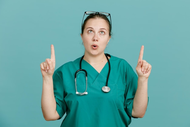 Jonge vrouw verpleegster in medisch uniform met stethoscoop om nek kijken verbaasd omhoog wijzend met wijsvingers over blauwe achtergrond