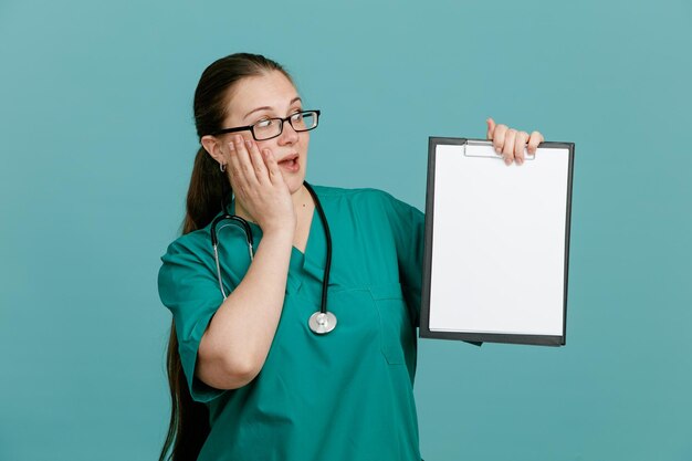 Jonge vrouw verpleegster in medisch uniform met stethoscoop om nek met klembord kijken verbaasd en verrast over blauwe achtergrond