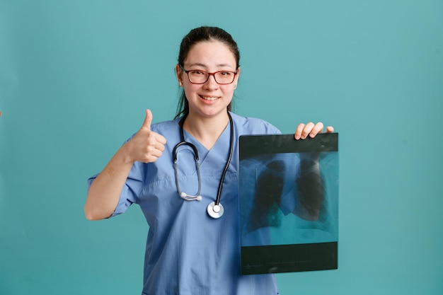 Jonge vrouw verpleegster in medisch uniform met stethoscoop om nek met long xray kijken camera met blij gezicht glimlachend duim opdagen staande over blauwe achtergrond