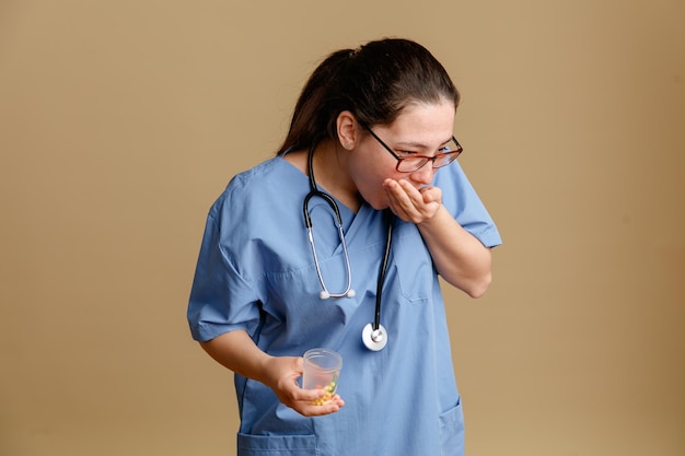 Jonge vrouw verpleegster in medisch uniform met stethoscoop om nek met pot met pillen die pillen innemen die over bruine achtergrond staan