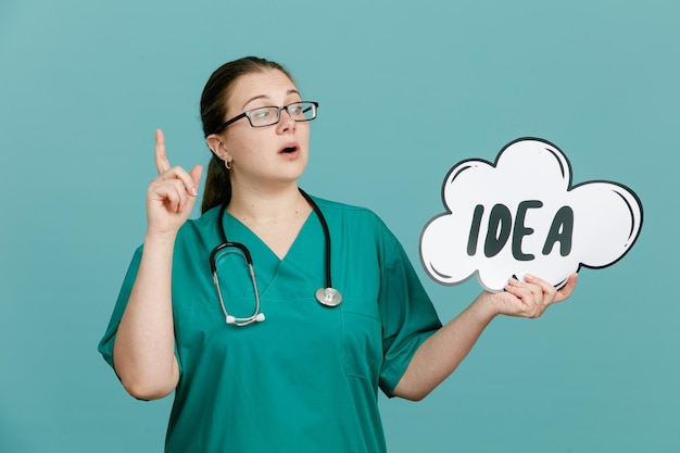 Jonge vrouw verpleegster in medisch uniform met stethoscoop om nek met zeepbel toespraak met woord idee op zoek zelfverzekerd tonen wijsvinger permanent over blauwe achtergrond