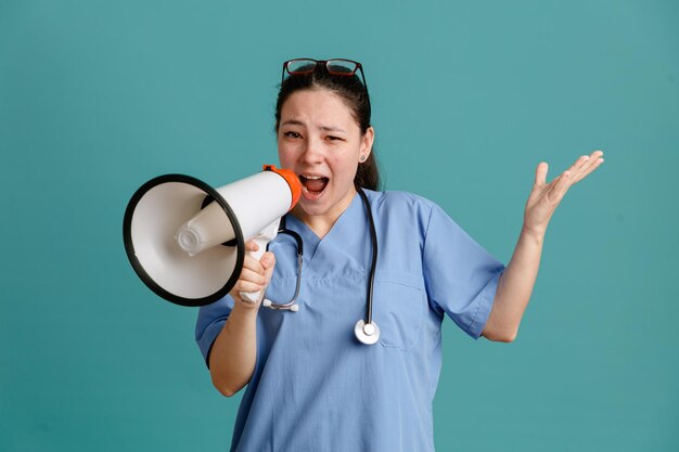 Jonge vrouw verpleegster in medisch uniform met stethoscoop rond nek schreeuwen in megafoon verward arm opheffen in ongenoegen over blauwe achtergrond