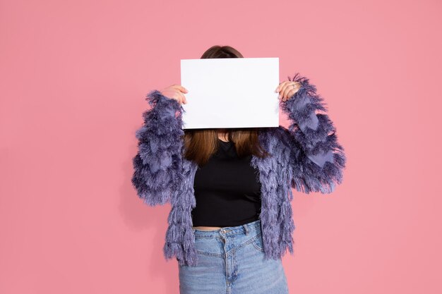 Jonge vrouw verstopt achter een wit blanco vel papier voor een inscriptie. Op roze achtergrond in studio. Onherkenbaar persoon.