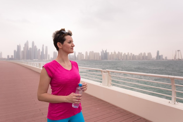 jonge vrouw viert een succesvolle training op de promenade aan zee met een fles water en haar handen in de lucht met een grote stad op de achtergrond