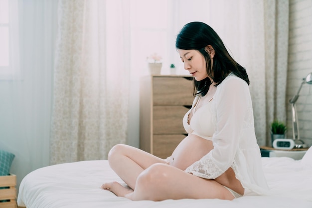 jonge vrouw voelt zich met handen die de babybult aanraken terwijl ze alleen in de slaapkamer rust. Aziatische Japanse zwangere dame zit op bed en lacht naar haar naakte buik. gelukkig ouderschap moeder met blote buik