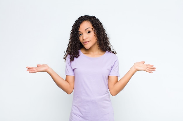 Jonge vrouw voelt zich verward en verward, onzeker over het juiste antwoord of de juiste beslissing, en probeert een keuze te maken over een witte muur