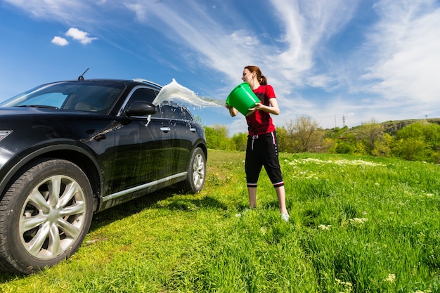 Jonge vrouw wassen zwarte luxevoertuig in met gras begroeide groene veld op heldere zonnige dag met blauwe lucht, emmer water op de zijkant van de auto gooien