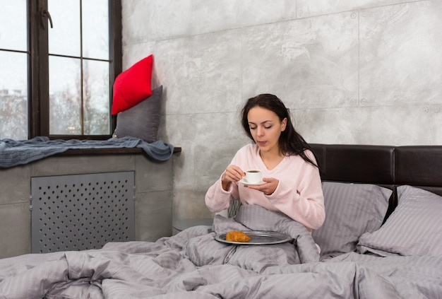 Jonge vrouw werd net wakker, met een kopje koffie en ontbijt op bed, gekleed in pyjama