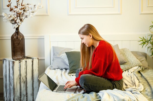 Jonge vrouw werkt op een computer, zittend op een bed, op afstand werkend. Een meisje met lang haar in een rode trui en spijkerbroek werkt thuis.