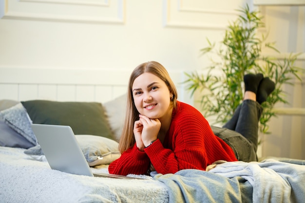 Jonge vrouw werkt op een computer, zittend op een bed, op afstand werkend. Een meisje met lang haar in een rode trui en spijkerbroek werkt thuis.