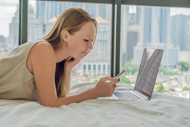 Jonge vrouw werkt op een laptop in zijn bed op een achtergrond van een panoramisch raam met uitzicht op de