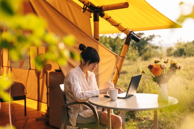 Jonge vrouw werkt op een laptop terwijl ze ontspannen op een stoel bij het kampvuur zit, reist met een tent, concept van afstandswerk en ontsnapping naar de natuur.