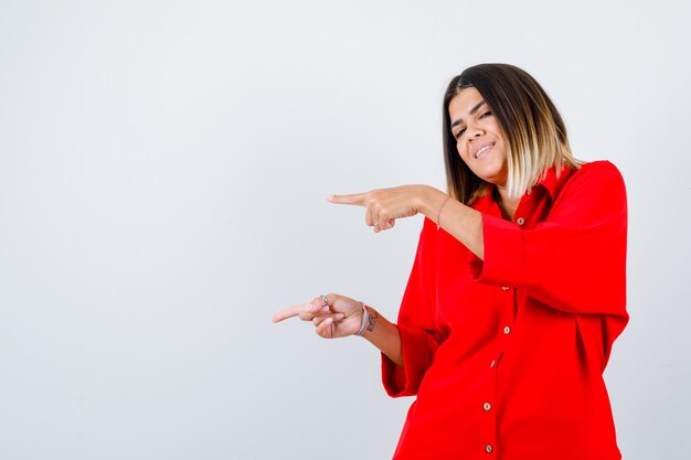 Jonge vrouw wijst naar de linkerkant in een rood oversized shirt en ziet er gelukkig uit. vooraanzicht.