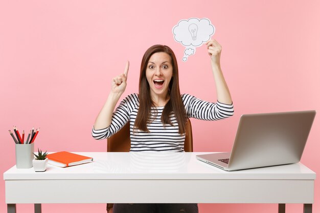 Jonge vrouw wijzende vinger omhoog houden Say cloud tekstballon met gloeilamp sit werk op wit bureau met pc laptop geïsoleerd op pastel roze achtergrond. Prestatie zakelijke carrière concept. Ruimte kopiëren.