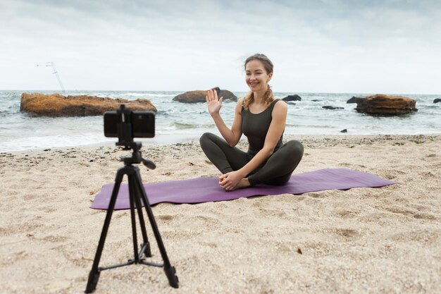 Jonge vrouw yogaleraar leidt online les op strand Sportvrouw zendt uit naar camera van smartphone die op statief staat Bloggen vlog Gezonde levensstijl