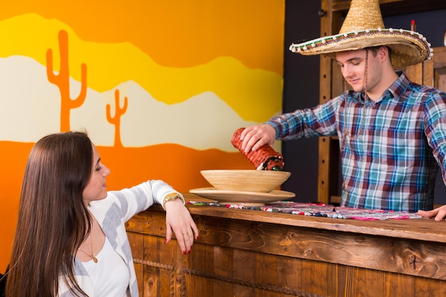 Jonge vrouw zit aan de bar terwijl barman in sombrero en een geruit hemd haar een drankje schenkt in een Mexicaanse bar