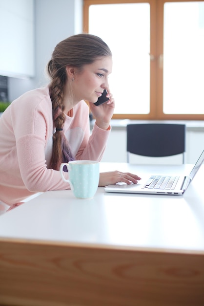 Jonge vrouw zit aan de keukentafel met een laptop en praat op een mobiele telefoon