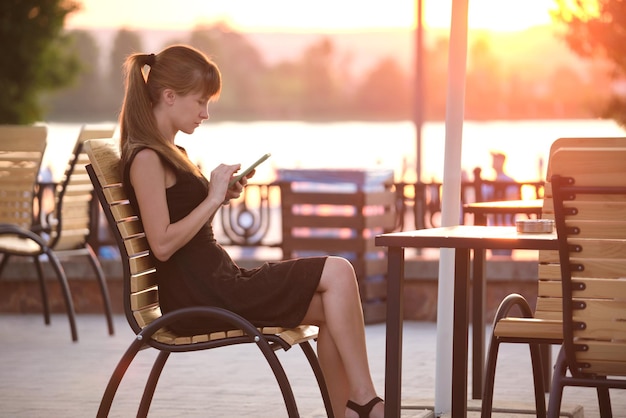 Jonge vrouw zit aan straat café tafel haar mobiel buiten te bladeren op warme zomeravond. Communicatie en mobiel verbindingsconcept.