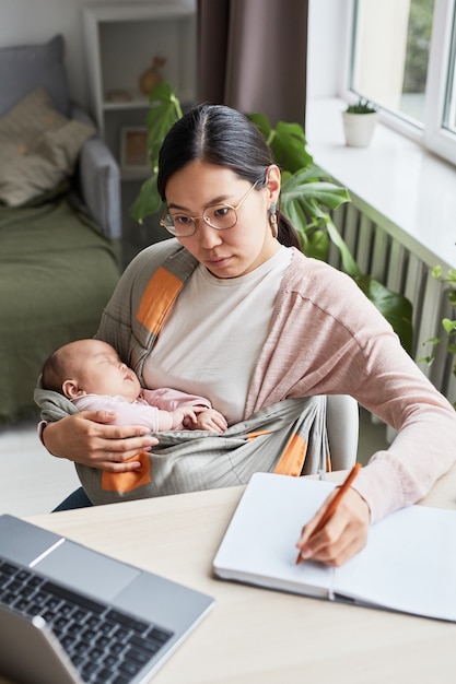 Jonge vrouw zit aan tafel met haar baby op handen en studeert thuis tijdens haar zwangerschapsverlof