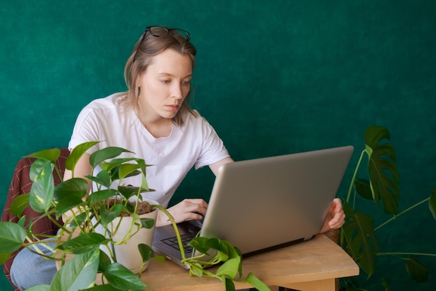 Jonge vrouw zit aan tafel met laptop thuis naast huisplanten in potten