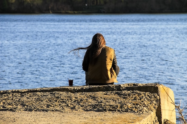 Jonge vrouw zit alleen op de pier met koffiekopje. Achteraanzicht