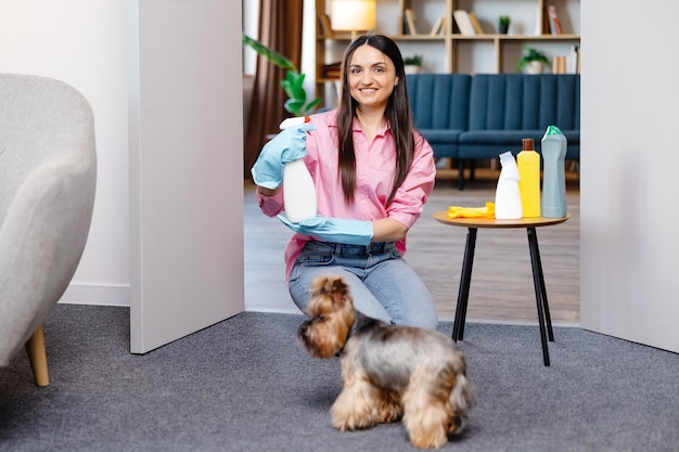 Jonge vrouw zit bij de tafel met schoonmaakproducten thuis en haar hondje rent naast haar