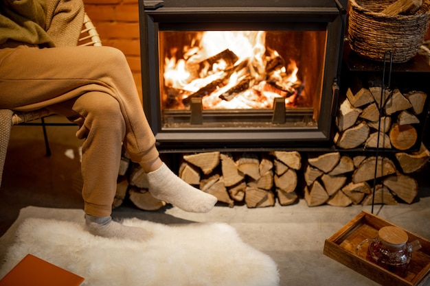 Jonge vrouw zit bij een brandende open haard, ontspannen met een warm drankje in een gezellig interieur in loftstijl tijdens een wintertijd. Bijgesneden afbeelding zonder gezicht gericht op een vlam