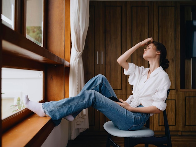 Jonge vrouw zit bij het raam met telefoon in de hand glimlacht online kijkend voorjaarsstemming thuis eenvoudige kleren thuis gezellige sfeer esthetische levensstijl