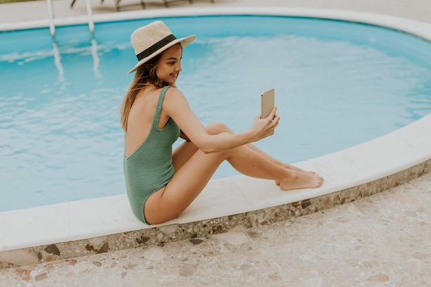 Jonge vrouw zit bij het zwembad en neemt selfie foto met mobiele telefoon in de achtertuin van het huis