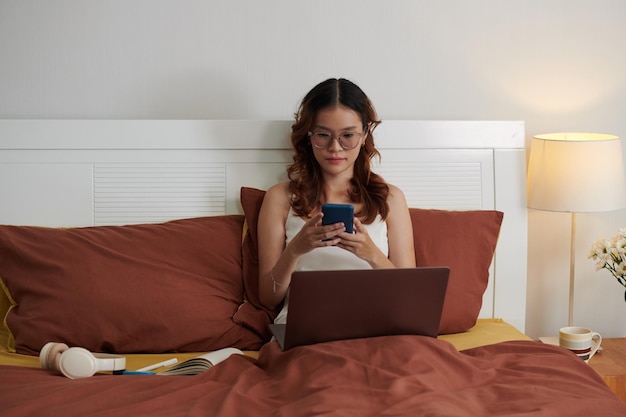 Jonge vrouw zit in bed met laptop en sms't collega's