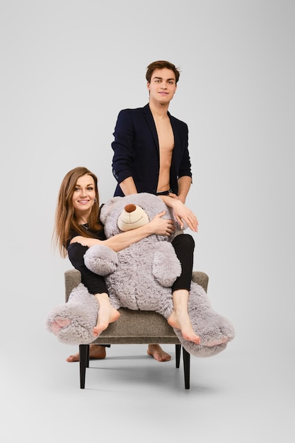Jonge vrouw zit in de fauteuil en houdt grote teddybeer vast en man staat naast haar