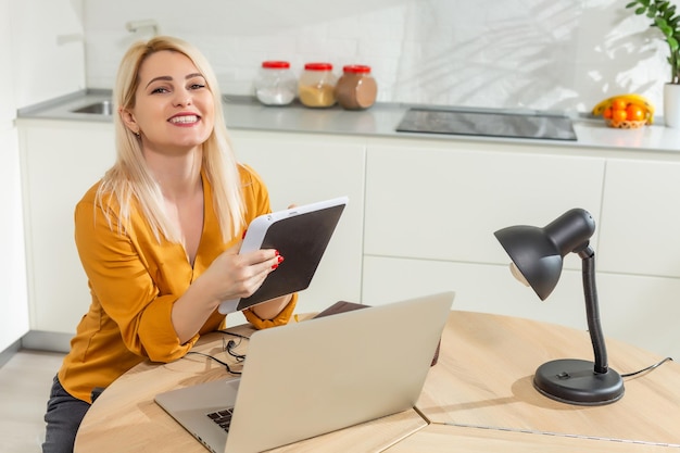 Jonge vrouw zit in de keuken en werkt op laptop.