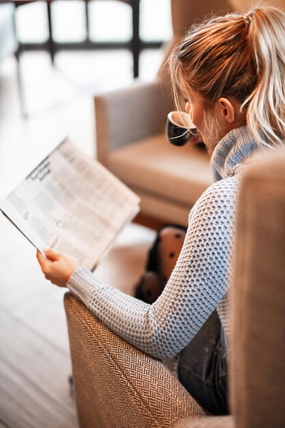 Jonge vrouw zit in een fauteuil en leest de krant terwijl ze koffie drinkt in een café